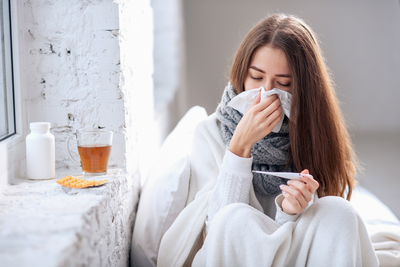 young woman with the flu wrapped up in blanket, blowing nose, and looking at thermometer