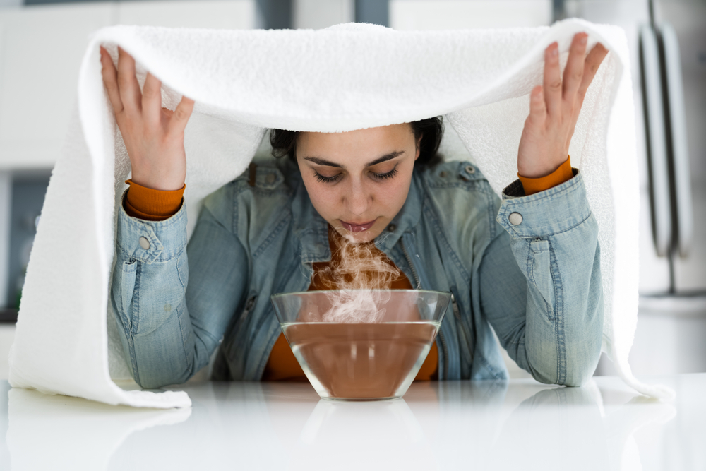 woman breathing in steam from bowl of hot water with white towel above her head